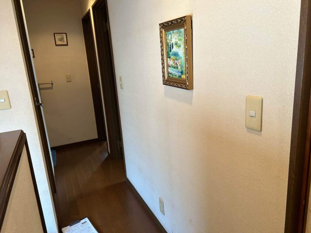 クロス貼り替え前の2階廊下の様子です。 経年によるクロスの傷みや、全体的なトーンの暗さが見受けられました。今回のリフォームでは、新しい量産品クロスへの貼り替えを行うとともに、デザインのアクセントとして壁にトリム（帯状の壁紙）をコの字型に施工し、明るく整った空間へと一新いたします。