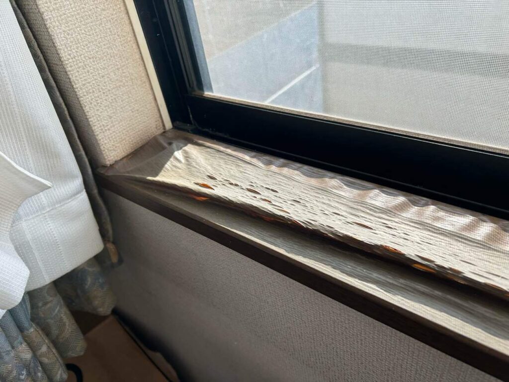 2階のお部屋にある窓枠の施工前の状態です。 結露や紫外線などの影響により、窓枠下部の表面シートが経年劣化で波打ち、剥がれが生じていました。今回の工事では、傷みが見受けられる窓枠下部のみにウォールナット柄のフィルム（ダイノックシート）を張り、新品同様の美観を取り戻します。