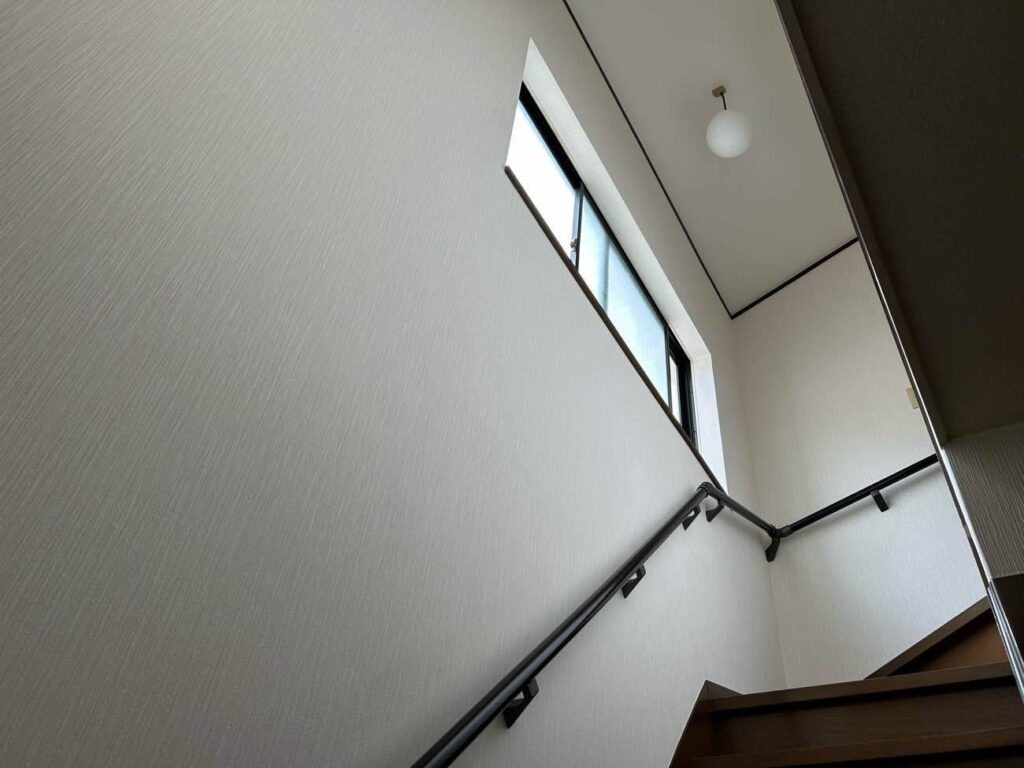 新しくLIXIL製の手すり（クリエダーク色）を設置し、壁と天井のクロス貼り替えが完了した階段の様子です。壁の内部に合板でしっかりと補強下地を造作した上で手すりを固定しているため、安全に体重をかけて昇降いただけます。明るいトーンのクロスに張り替えたことで空間全体が明るくなり、落ち着いた色合いの手すりと調和した安全な階段へと仕上がりました。