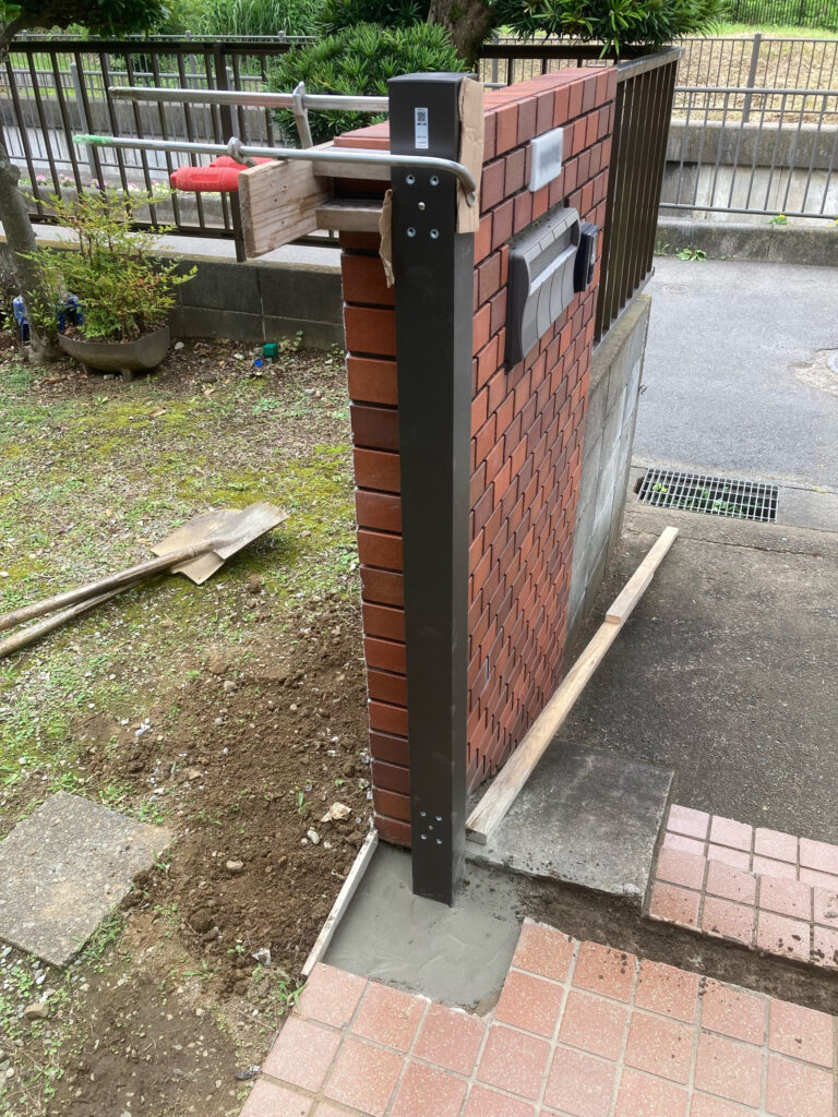 既存の古い門扉を撤去し、新しい門扉の柱を取り付けている施工中の様子です。あらかじめ古い柱を抜き取って空けておいた穴に、新しいアルミ製の柱を垂直に立て、モルタルを流し込んでしっかりと固定していきます。門扉は毎日の開閉で負荷がかかるため、少しの傾きもないよう水平器などを用いてミリ単位で正確に位置を調整しながら作業を進めます。モルタルが完全に硬化するまでしっかりと養生期間を設け、土台を作ります。